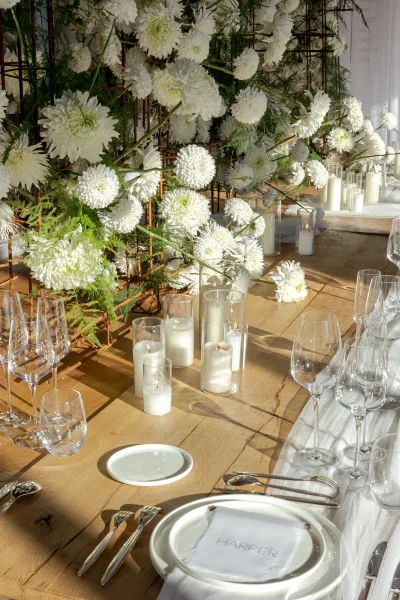 Reception tablescape with a wedding table centerpiece of white florals and greenery, pillar candles in glass on a wooden table with draping behind