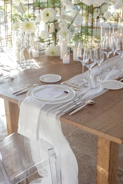 Reception tablescape with wedding table setting on a wood dining table, white florals and greenery, pillar candles, and sunlit windows behind