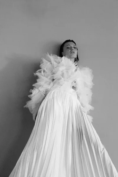 Bridal portrait of a bride in a white wedding dress with ruffled tulle sleeves and pleated skirt, wearing earrings against a plain wall