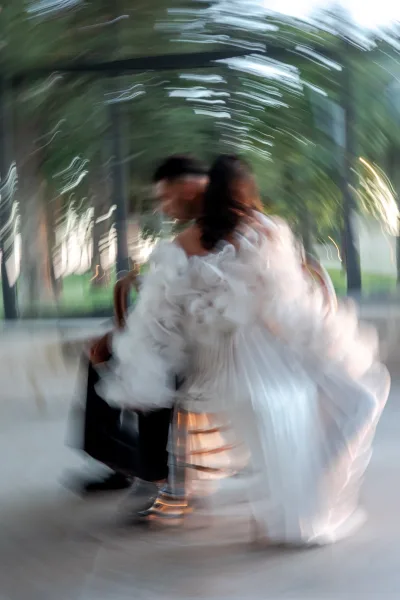 Couple portrait of bride and groom sitting on a ledge, her strapless tulle skirt draping as trees and a glass wall frame them