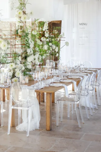 Reception tablescape with long banquet table setup on wood farm tables, white runner, acrylic chairs, white florals, candles, and place cards indoors