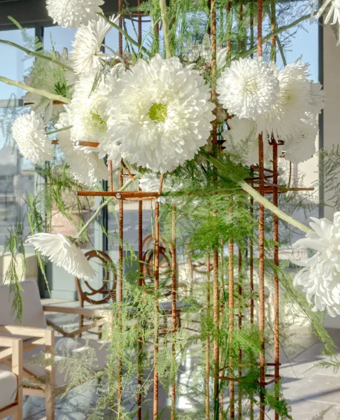 Wedding floral installation of white chrysanthemums and gerbera daisies with greenery on a metal grid stand, lit by daylight near glass windows