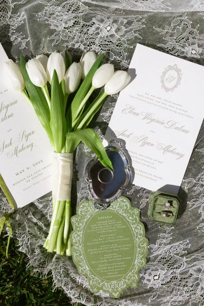Wedding invitation suite with green and white wedding invitations, styled with a white tulip bouquet on lace fabric over a grass lawn.