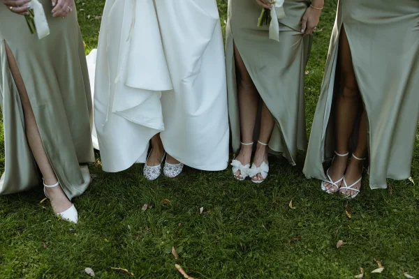 Bridesmaid dresses in sage green satin with thigh-high slits, white ribbon-wrapped bouquets, and heels on a grass lawn
