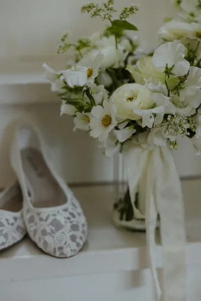 Bridal bouquet of white wedding bouquet blooms and greenery with long ribbon wrap in a glass jar on painted steps beside lace flats