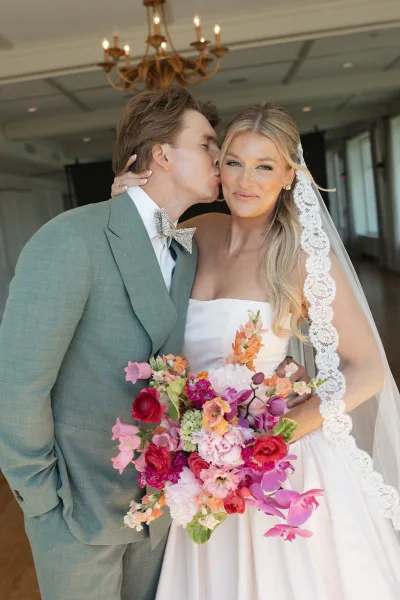 Couple portrait with groom kissing bride, her vibrant bouquet standing out in a chandelier-lit indoor room with tall windows.