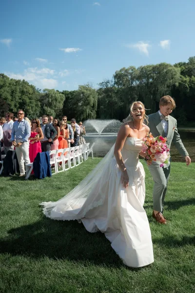Ceremony recessional as the couple make their wedding aisle exit, bride carrying a colorful bouquet, with a lakeside fountain and trees beyond.