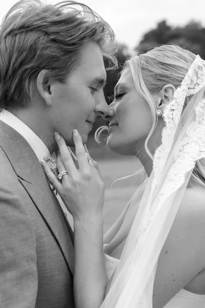 A black-and-white couple portrait of newlyweds touching noses with eyes closed, her bridal veil gliding over his shoulder in a tender side profile
