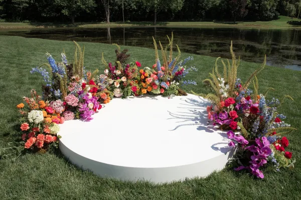 Wedding altar on a circular ceremony platform, edged with lush orchids, set on a grass lawn beside a pond and trees.