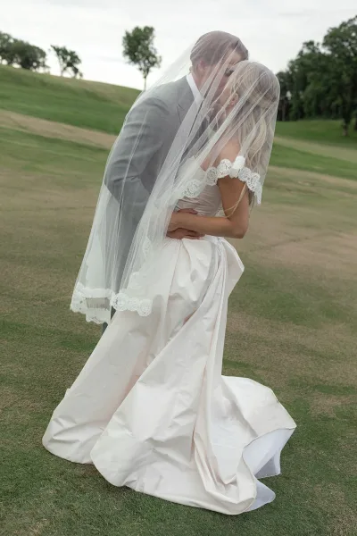 Wedding kiss portrait under a shared veil, the groom holding the bride’s waist as soft natural light frames their faces.