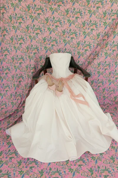 Strapless wedding dress with a ball gown silhouette and ribbon sash, set against a pink floral patterned backdrop.