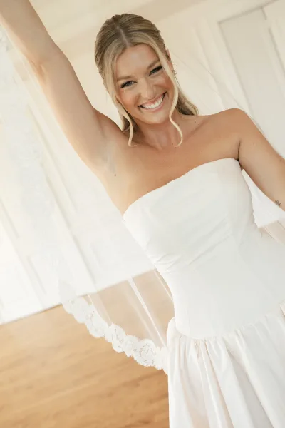 Bridal portrait of a bride with veil, lace veil draped over her shoulders before a white paneled wall and warm hardwood floor.