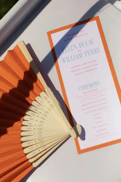 Wedding program, an order of service card with an orange border and pink-blue text, paired with a paper hand fan on a white tabletop by a grass lawn.