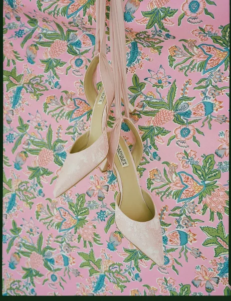 Bridal shoes—pink wedding shoes with delicate ribbon ties—rest on pink floral fabric, creating a soft, romantic accessory vignette.
