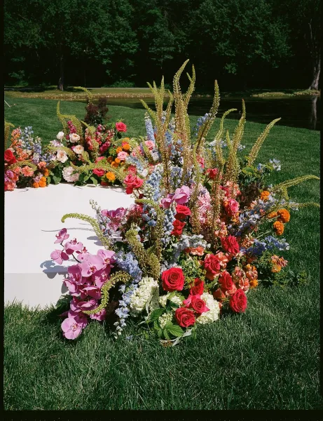 Ceremony floral installation with ground ceremony flowers, lush with roses, on a grass lawn beside a pond, framed by trees and forest.