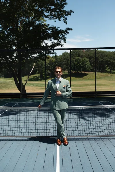 Groom portrait in a sage green groom suit with a bow tie, standing on a tennis court by the net under a clear sky.