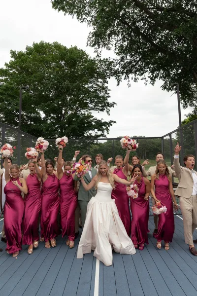 Wedding party photo capturing a bridal party group shot with magenta bridesmaid dresses on a blue tennis court under a cloudy sky.