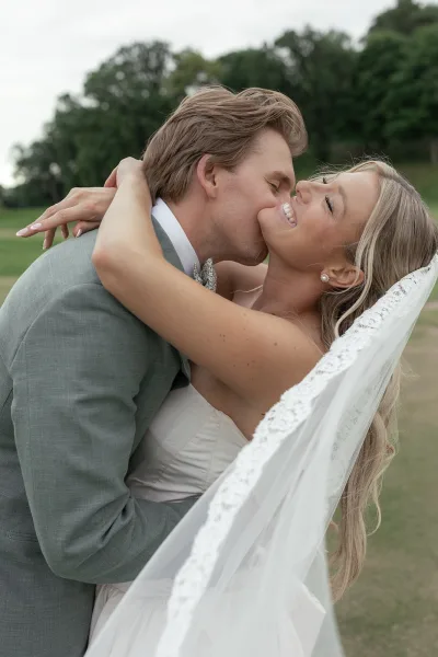 Wedding kiss portrait of a groom kissing his bride as she smiles, her lace-edged veil draping over his shoulder in a tender close-up.