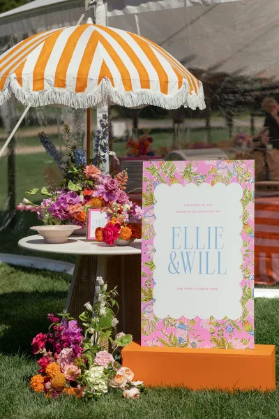 Wedding welcome sign anchors an entrance sign display under a clear tent, paired with a striped umbrella on a garden lawn.