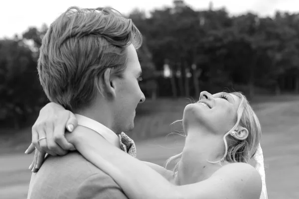 Couple portrait as the bride and groom embrace, her bridal veil catching light while they smile close together outdoors beneath leafy trees.