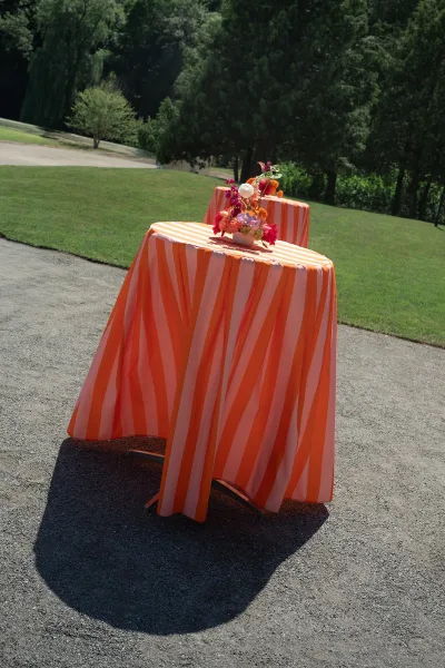Cocktail table decor at an outdoor cocktail hour, draped in a striped tablecloth on a grass lawn with trees and a nearby gravel path.