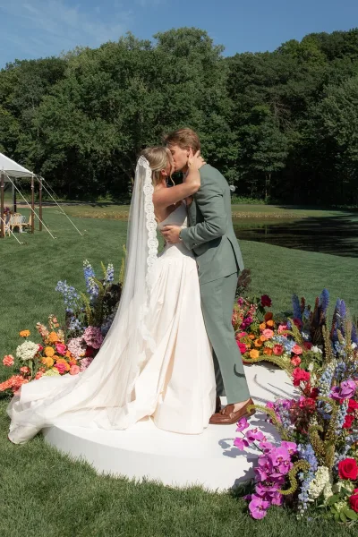 Wedding first kiss in an outdoor ceremony moment, the couple embracing amid wildflower florals, her veil catching light by a pond.