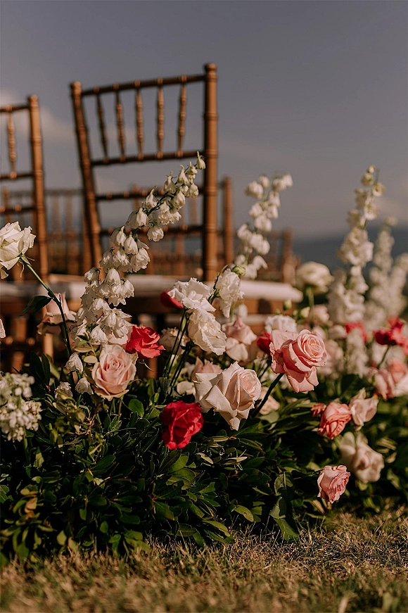 Ceremony aisle florals with roses and white flower spikes in grounded meadow-style arrangements beside chiavari chairs on a coastal lawn