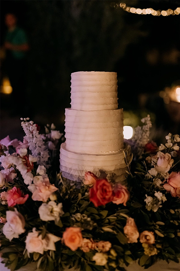 Wedding cake with three tier white buttercream frosting, wrapped in rose and greenery garland on a candlelit table with bokeh string lights
