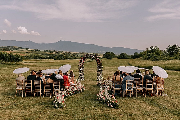 Ceremony setup for an outdoor wedding ceremony with a rose floral arch and aisle florals, framed by white parasols and a mountain view