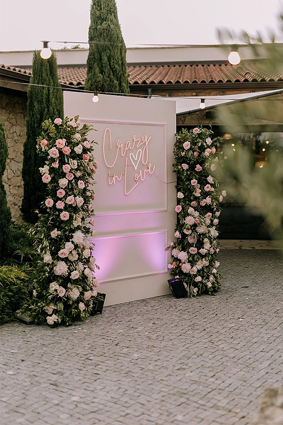 Wedding neon sign reading crazy in love on a white panel backdrop with a rose and greenery arch, in a courtyard with string lights