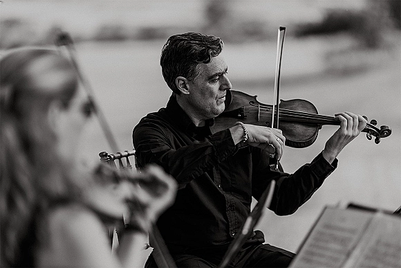 Wedding musician playing violin with bow near sheet music on a chair, set in an outdoor ceremony with softly blurred performer behind