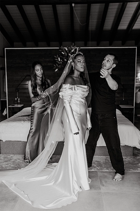 Bridal getting ready as a bridesmaid helps the bride putting on veil, showcasing a long satin off-the-shoulder gown in a cozy bedroom