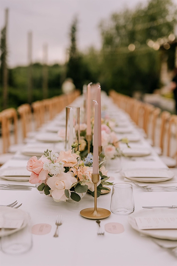 Reception tablescape with white linen, blush taper candles in brass candlesticks, low rose greenery centerpieces under garden string lights.