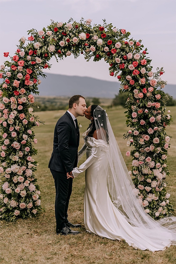 Wedding kiss portrait of bride and groom kissing under a rose-covered floral arch, her veil flowing in a meadow with mountains behind