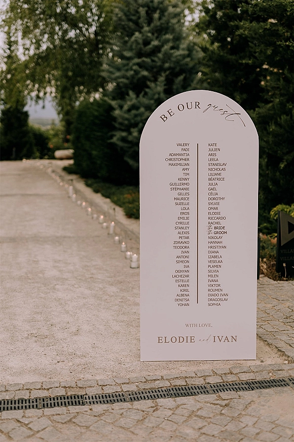 Wedding seating chart on an arched seating chart sign with calligraphy guest list and glass candle holders along a gravel garden pathway