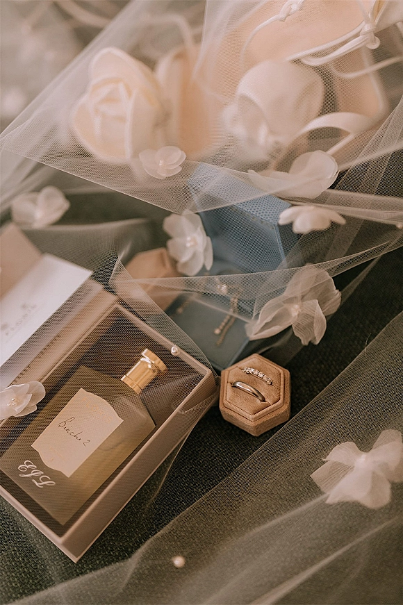 Wedding rings displayed in a ring box with a perfume bottle and ribbon on dark fabric, with veil appliques and pearl beads nearby