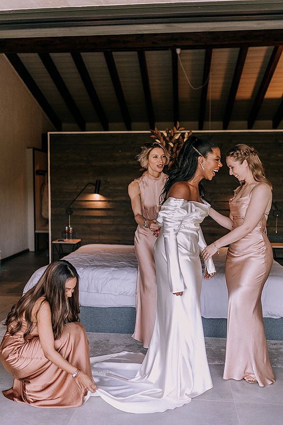 Bride getting ready as bridesmaids help adjust her off-the-shoulder satin wedding dress train in a hotel bedroom with wooden headboard