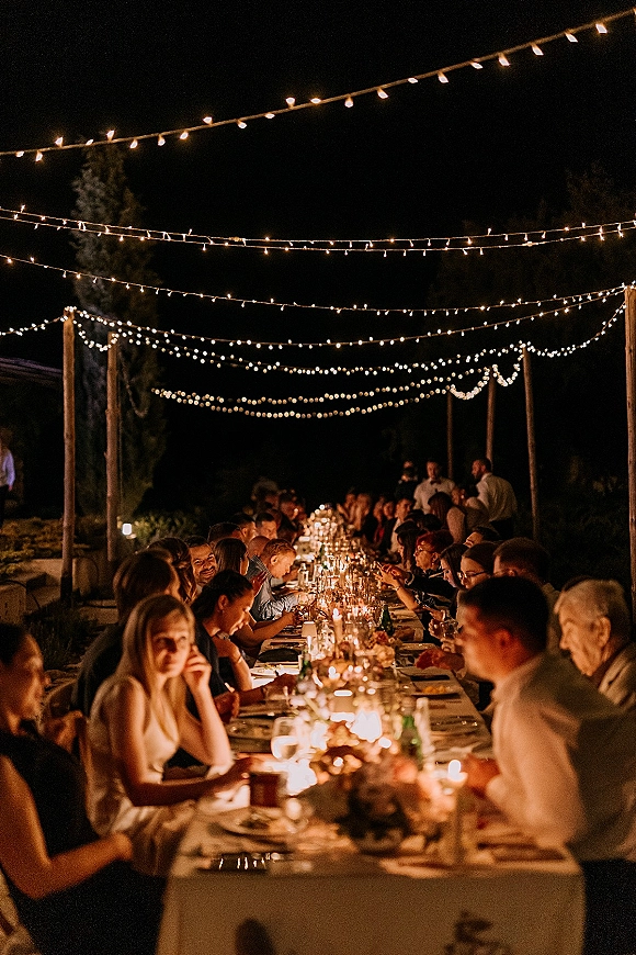 Wedding reception dinner at a long banquet table wedding under string lights, with candles and floral runner on an outdoor patio at night