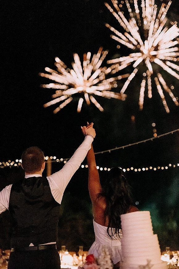 Wedding fireworks moment as bride and groom hold hands from behind, watching bursts above string lights by the cake at outdoor reception
