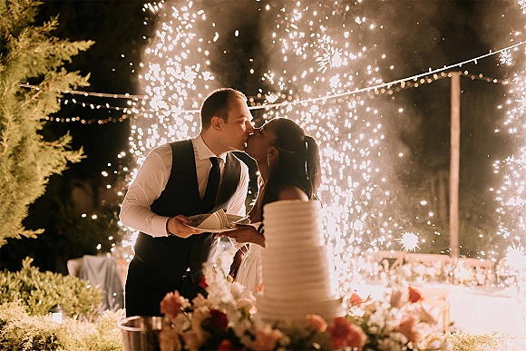 Wedding cake cutting as the couple kisses by a tiered white cake under string lights and sparklers at an outdoor night reception