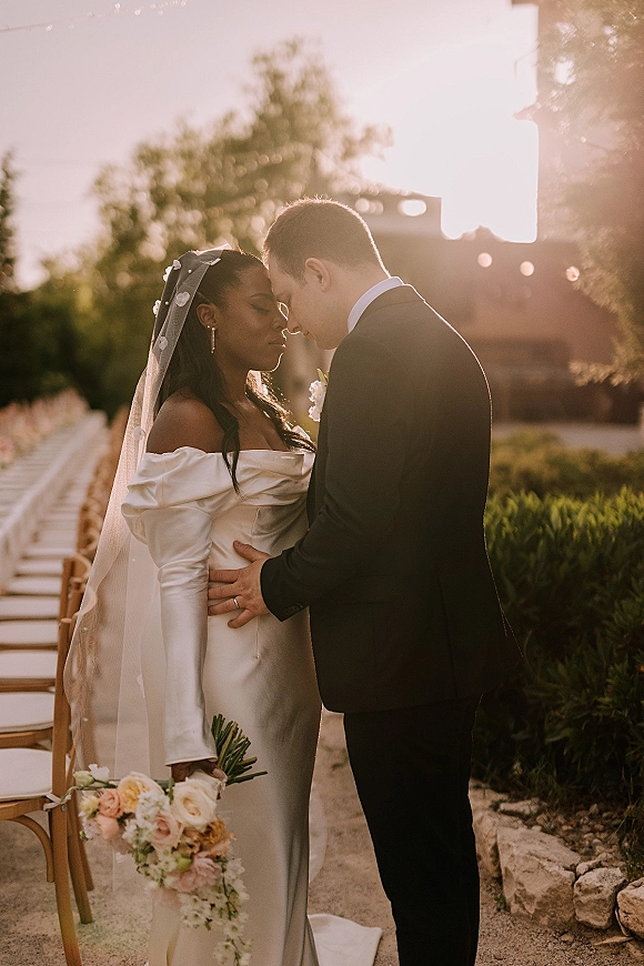 Couple portrait of bride and groom forehead touch, backlit by sunlight, bride in veil holding bouquet on garden aisle near stone building
