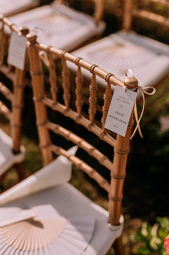 Wedding escort card tied with a ribbon bow to a wooden chiavari ceremony chair tag, with white cushion and paper fan on grass