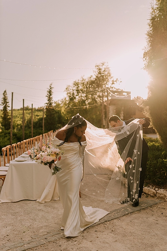 Couple portrait of bride and groom with veil, bride holding bouquet beside a long alfresco reception table under string lights at sunset
