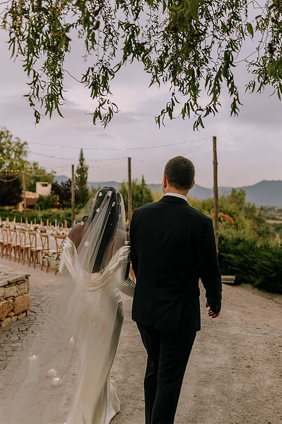 Newlywed couple walking away hand in hand, bride’s veil and satin gown beside string lights over a long terrace reception table at dusk