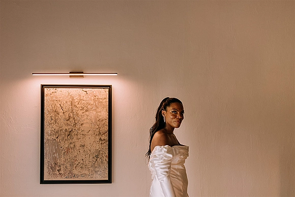 Bridal portrait of a bride in an off the shoulder wedding dress, looking over her shoulder by a neutral wall with framed abstract art and sconce light