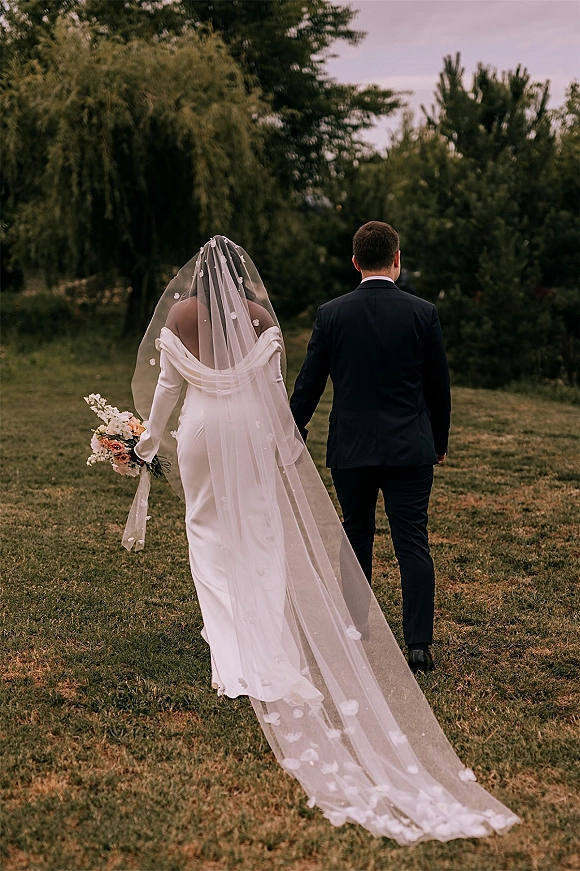 Couple walking away hand in hand, newlyweds walking hand in hand across a grassy lawn, bride’s long veil and bouquet trailing behind