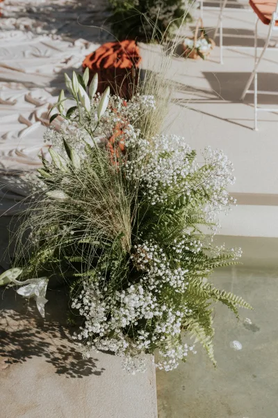 Ceremony aisle flowers with wedding aisle floral arrangements on stone pedestals, ferns, lilies, and baby's breath along sunlit patio steps