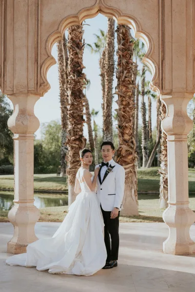 Couple portrait of bride and groom portrait under a stone archway, her long dress train flowing as he wears a white tuxedo jacket and boutonniere