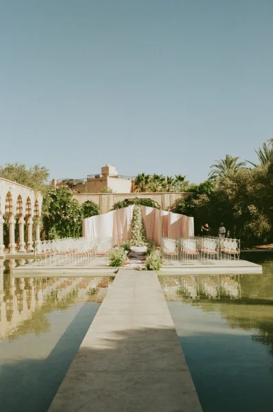 Ceremony setup with outdoor ceremony aisle leading to a white draped floral arch beside a reflecting pool in a palm-lined courtyard