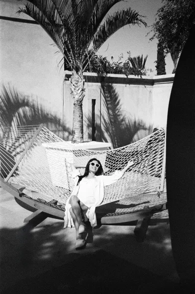 Bridal getting ready portrait of a bride in robe lounging in a hammock, wearing sunglasses on a sunlit patio with palm shadows